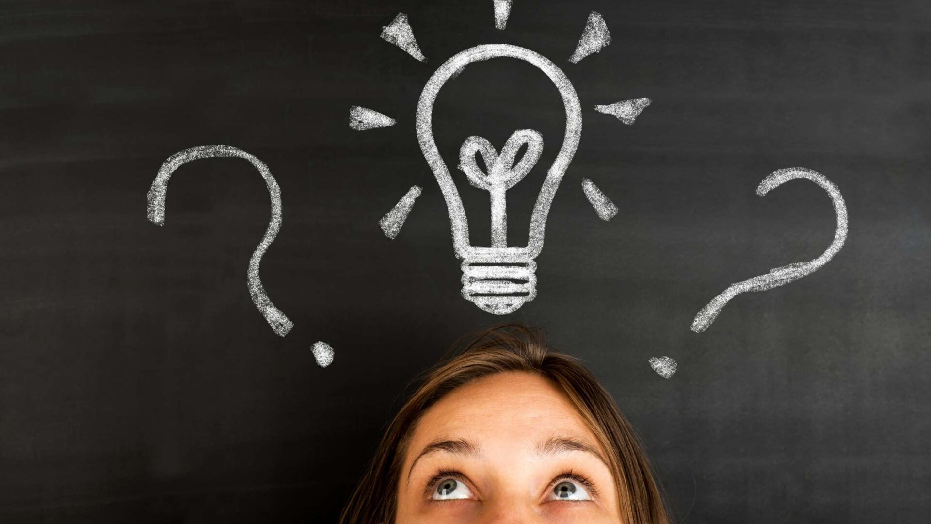 A woman looking up thoughtfully at a chalk-drawn lightbulb and question marks on a blackboard, symbolising brainstorming, ideas, and problem-solving.