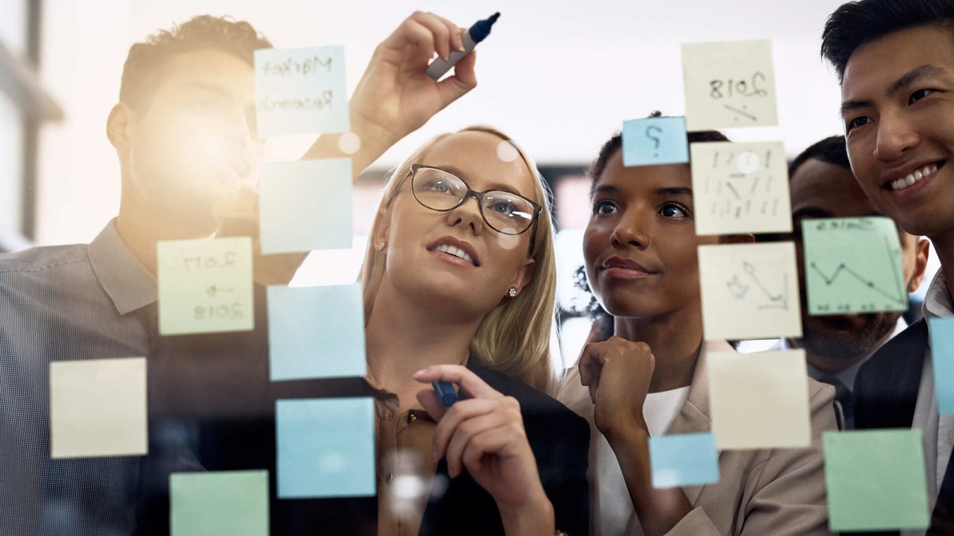 A diverse team of professionals collaborating in front of a glass board covered with sticky notes, writing and brainstorming ideas together in a bright, modern office setting.