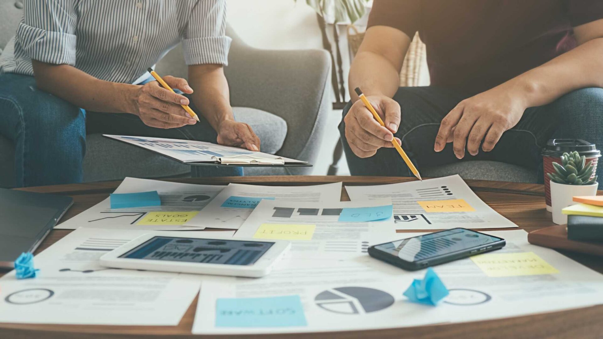 Two people who plan to start a new business at a wooden table covered with charts, graphs, sticky notes, and digital devices, including a smartphone and tablet, with a coffee cup and a small potted plant nearby.