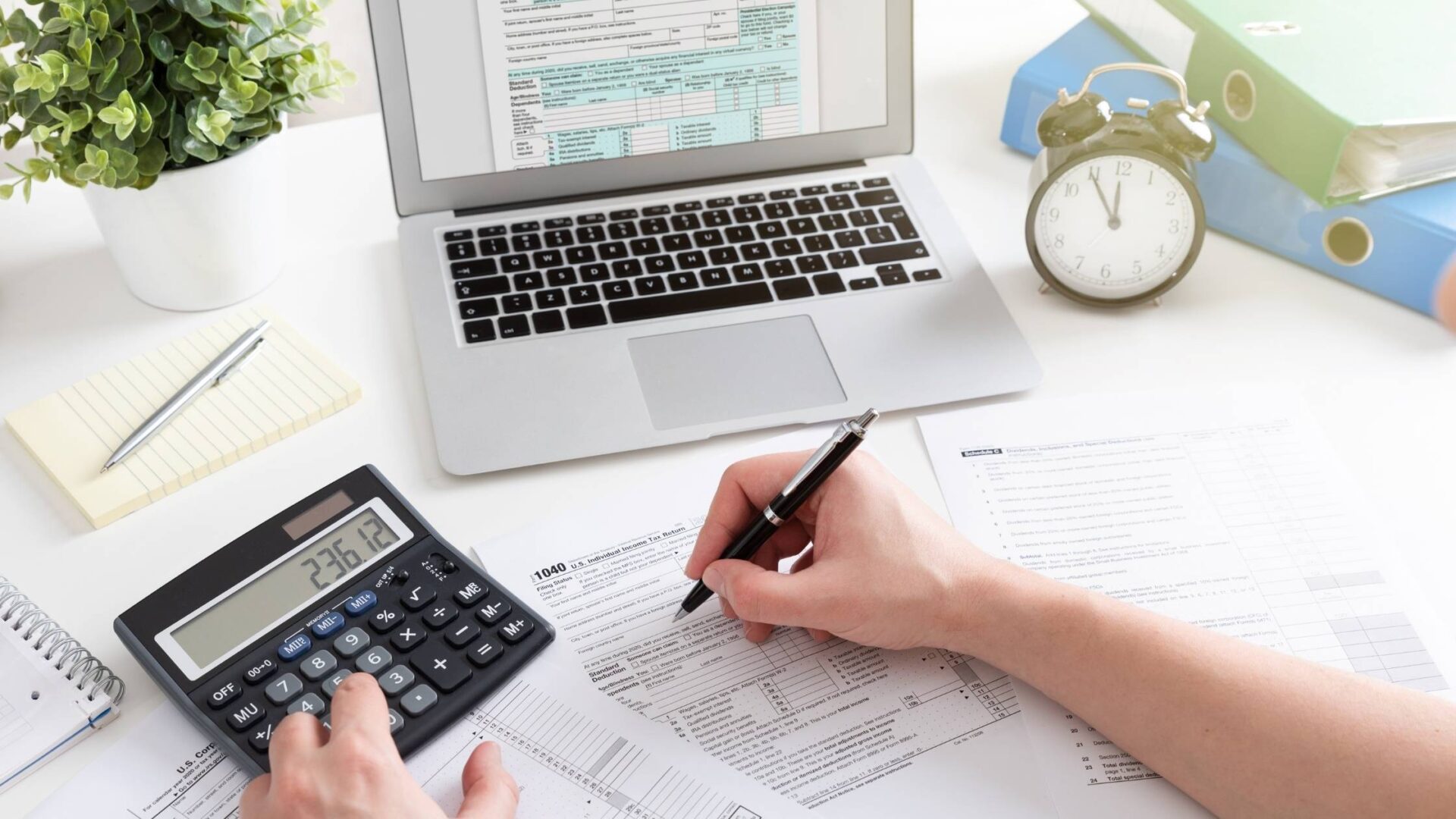 A bookkeeper calculating finances with a laptop, documents, and a calculator on a desk.